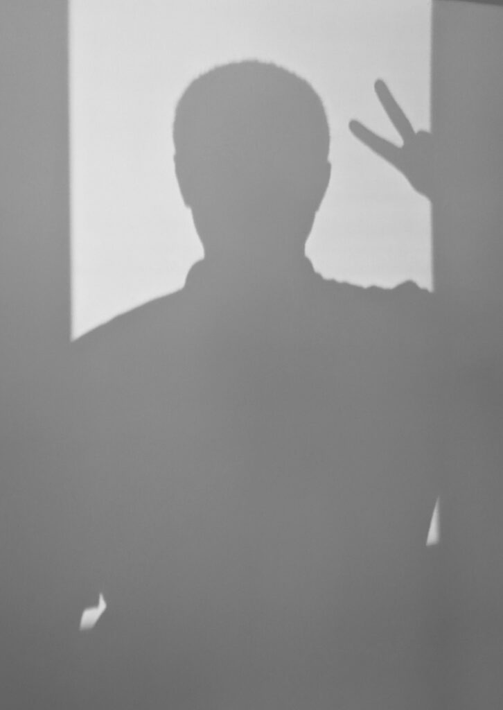 Shadow silhouette of a person showing a V-sign against a white wall, symbolizing peace or victory.